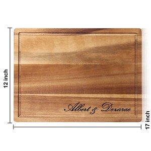 May include: A rectangular wooden cutting board with a natural wood grain finish. The board is 17 inches wide and 12 inches long. It has a groove around the edge and is engraved with the text "Albert & Dexarae".