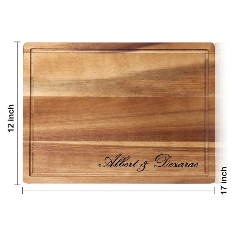 May include: A rectangular wooden cutting board with a natural wood grain finish. The board is 17 inches wide and 12 inches long. It has a groove around the edge and is engraved with the text "Albert & Dexarae".