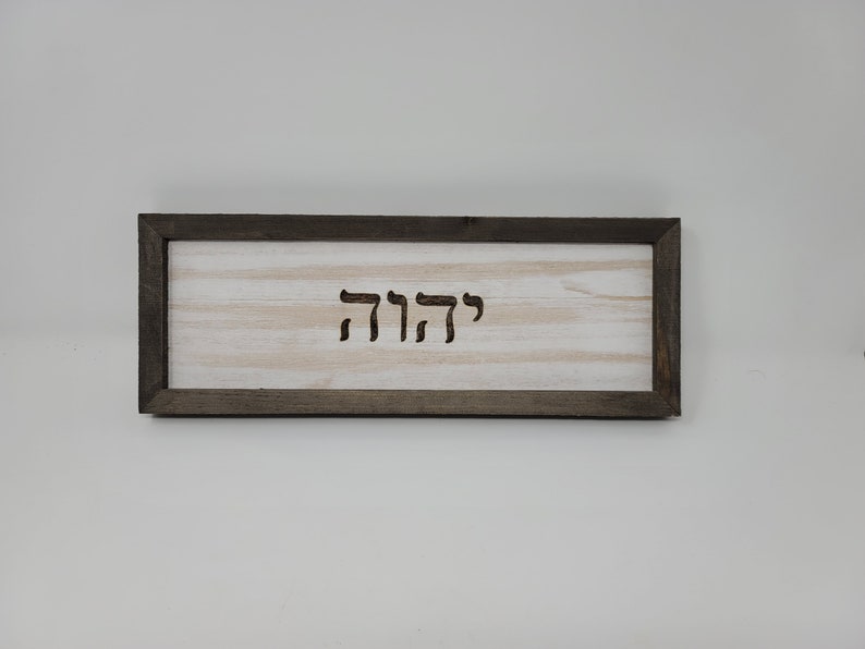 May include: A wooden sign with a dark brown frame and a white background. The sign has the Hebrew word "קדושה" carved into it.