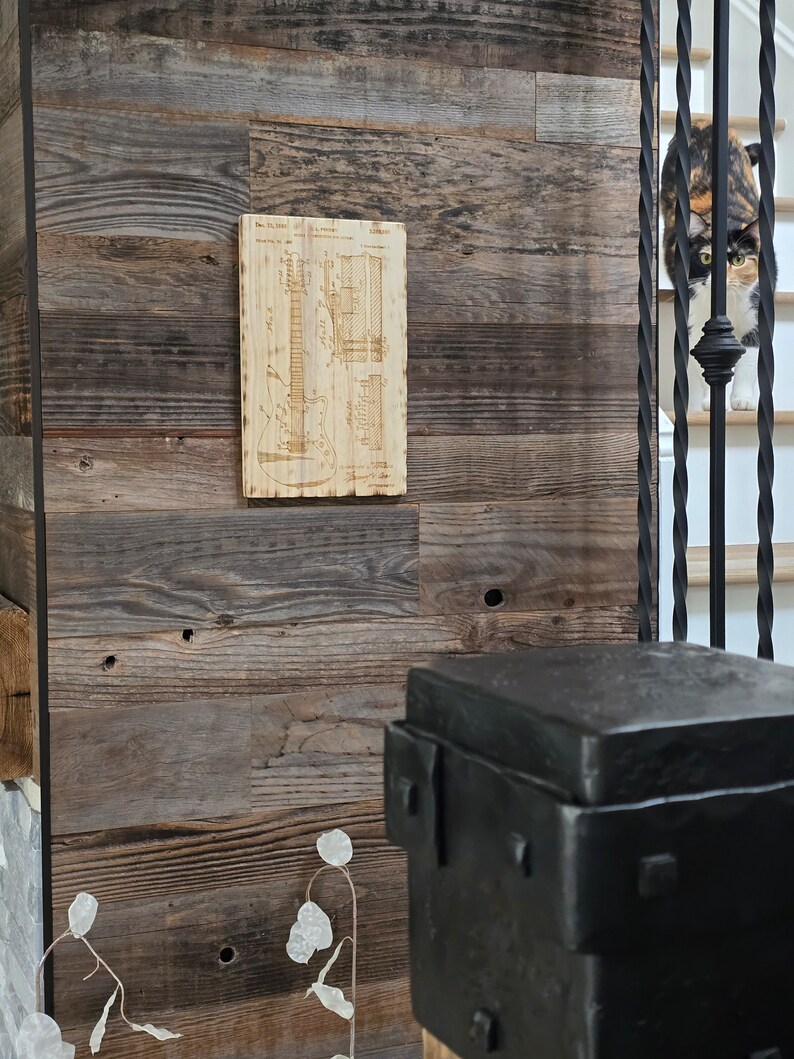 May include: A wood-panelled wall with a vintage guitar blueprint design printed on a wooden plaque. The plaque is mounted on the wall. A black metal stool is in the foreground.