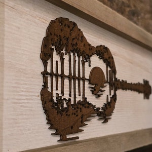 Puede incluir: Una pieza de arte de pared de madera con la silueta de una guitarra ac&uacute;stica con una puesta de sol y &aacute;rboles dentro del cuerpo de la guitarra.