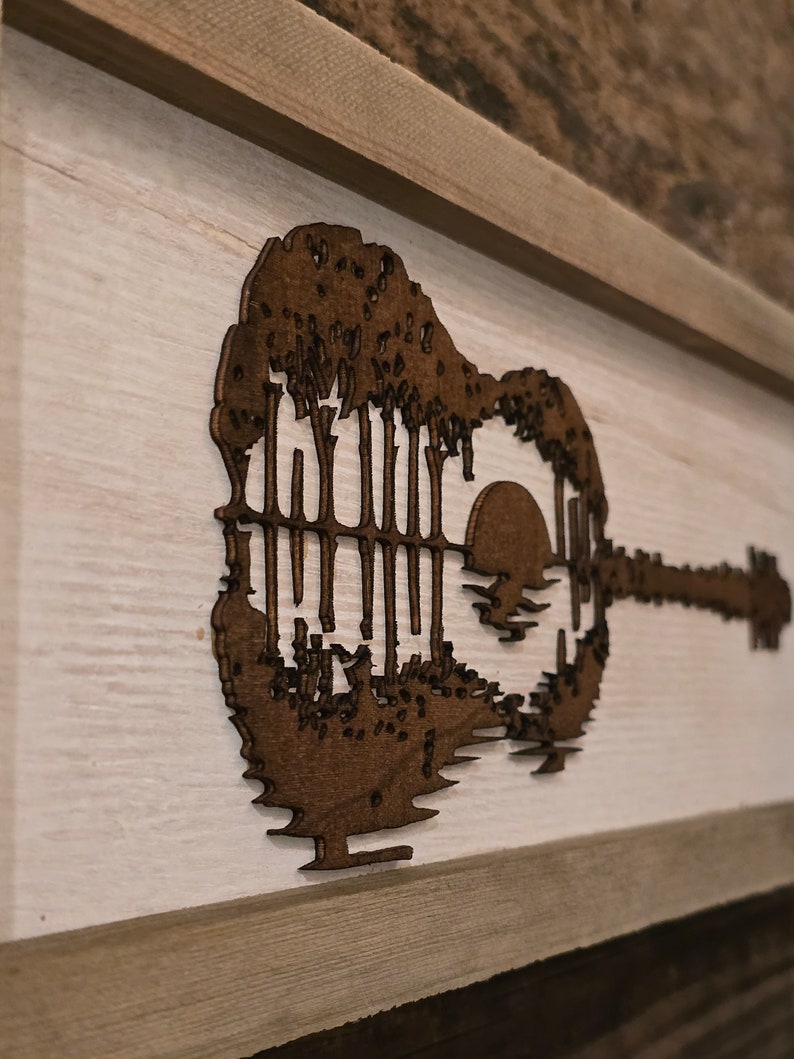 Puede incluir: Una pieza de arte de pared de madera con la silueta de una guitarra ac&uacute;stica con una puesta de sol y &aacute;rboles dentro del cuerpo de la guitarra.