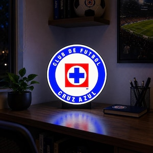 May include: A circular, illuminated sign featuring the Club de Futbol Cruz Azul logo, which consists of a blue ring with the text Club de Futbol Cruz Azul surrounding a white square containing a blue cross on a red background.