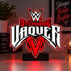 May include: A custom LED sign featuring the WWE logo at the top, the name STEPHANIE VAQUER in bold white lettering, and a large red V below, all mounted on a black rectangular base with a glowing red outline.
