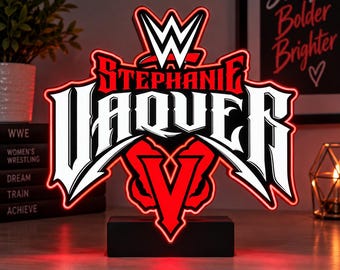 May include: A custom LED sign featuring the WWE logo at the top, the name STEPHANIE VAQUER in bold white lettering, and a large red V below, all mounted on a black rectangular base with a glowing red outline.
