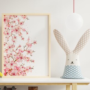 Puede incluir: Una pintura de acuarela enmarcada de una rama de cerezo rosa con fondo blanco. La pintura se exhibe en un estante blanco con un juguete de peluche de gato blanco, un juguete de peluche de monstruo amarillo, un juguete de peluche de conejo de madera y dos cestas de almacenamiento de tela con caras de oso.
