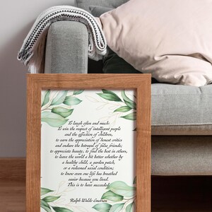 May include: A framed print with a wooden frame, featuring a quote by Ralph Waldo Emerson. The quote is in black text on a white background, with green botanical accents. The frame is brown and the print is displayed on a gray couch.