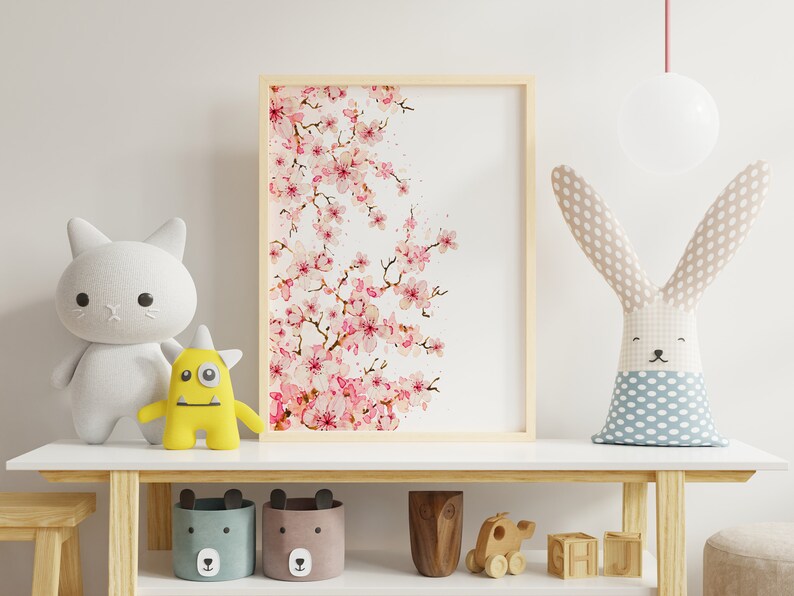 Puede incluir: Una pintura de acuarela enmarcada de una rama de cerezo rosa con fondo blanco. La pintura se exhibe en un estante blanco con un juguete de peluche de gato blanco, un juguete de peluche de monstruo amarillo, un juguete de peluche de conejo de madera y dos cestas de almacenamiento de tela con caras de oso.
