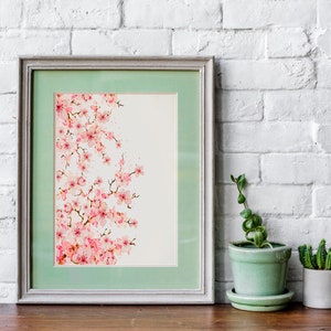 Puede incluir: Una pintura de acuarela enmarcada de una rama de cerezo rosa sobre un fondo blanco. El marco es de color gris claro con un passe-partout verde claro. La pintura est&aacute; sobre una mesa de madera con dos plantas en macetas.