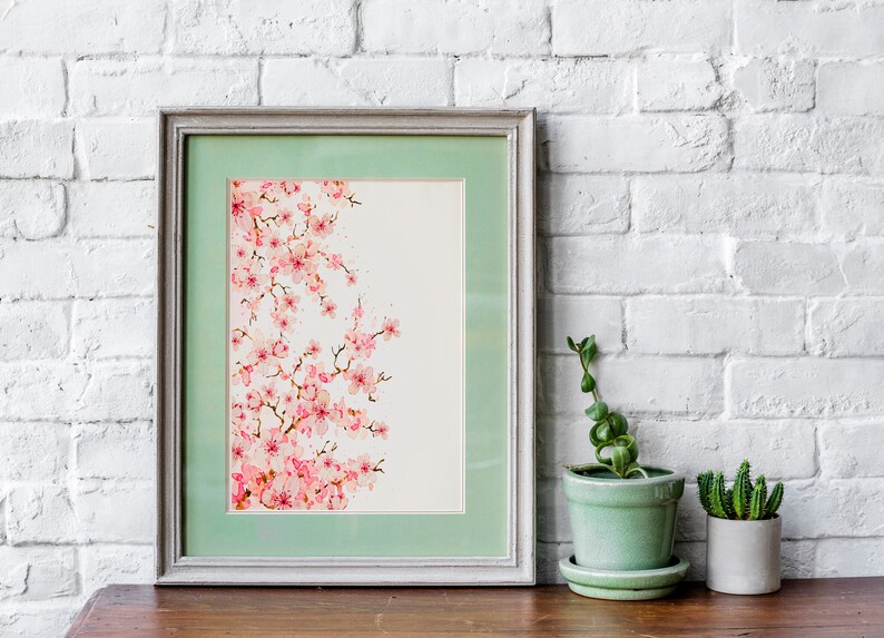 Puede incluir: Una pintura de acuarela enmarcada de una rama de cerezo rosa sobre un fondo blanco. El marco es de color gris claro con un passe-partout verde claro. La pintura est&aacute; sobre una mesa de madera con dos plantas en macetas.