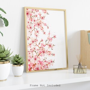 Puede incluir: Una pintura de acuarela de una rama de cerezo rosa sobre fondo blanco. La pintura est&aacute; enmarcada en un marco dorado y se exhibe en un estante blanco con plantas y un difusor.
