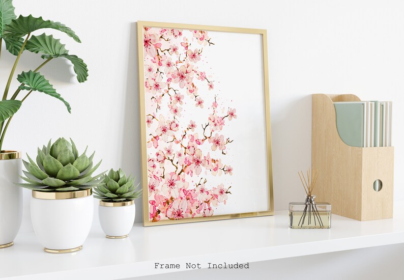 Puede incluir: Una pintura de acuarela de una rama de cerezo rosa sobre fondo blanco. La pintura est&aacute; enmarcada en un marco dorado y se exhibe en un estante blanco con plantas y un difusor.