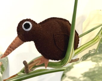 Puede incluir: Un kiwi de fieltro marrón hecho a mano con un pico largo y marrón y ojos con borde blanco. El ave está posada en un tallo de planta verde y blanco, mostrando su detallada artesanía y diseño natural. Un artículo decorativo encantador y caprichoso.