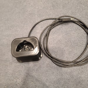 May include: A black charging stand with a cord for a wireless earbud case. The stand has a square base and a cutout for the case.
