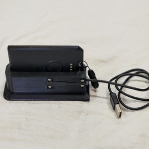 May include: A black electronic device in a black charging dock with a USB cable. The dock has a rectangular base and a slot to hold the device. The USB cable is coiled to the right of the dock.