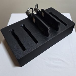 May include: A black rectangular organizer with four slots, holding two black keychains. The keychains have a metal ring and a rectangular tag. The organizer sits on a white surface.