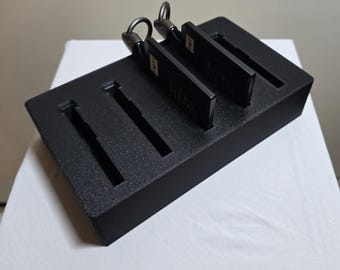 May include: A black rectangular organiser with four slots, holding two black keychains. The keychains have metal rings. The organiser sits on a white surface.