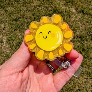 May include: A yellow glitter sun with a smiling face, attached to a clear plastic badge holder.
