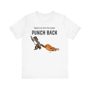 以下が含まれることがあります： 「WHEN LIFE GETS YOU DOWN PUNCH BACK」のテキストと、カウボーイの猿が人を引っ張っている漫画のイラストが描かれた白いTシャツ。イラストは茶色、黒、タンの色合いです。