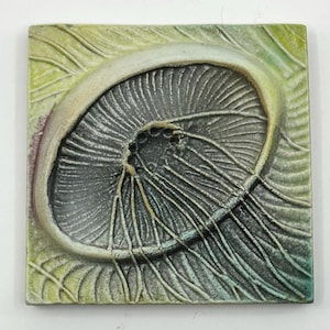 May include: Square ceramic tile with a detailed relief of a mushroom. The mushroom is gray with a white edge, set against a green and yellow background with leaf-like patterns. The tile has a textured surface.