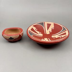 May include: Two pieces of pottery, a small bowl and a larger plate, both in a reddish-brown hue. The plate features a geometric design in black, tan, and white. The small bowl has a similar design around its rim.