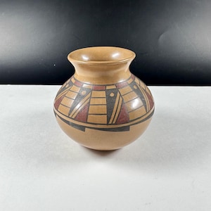 Op de afbeelding: Een kleine, ronde, lichtbruine aardewerken vaas met een brede opening. De vaas heeft een geometrisch ontwerp in zwart, rood en lichtbruin rond de body. Het ontwerp omvat driehoeken, rechthoeken en lijnen. De vaas is waarschijnlijk handgemaakt.