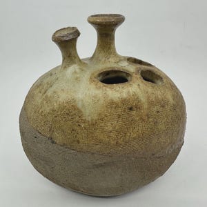 May include: A round, beige ceramic vase with a textured, cross-hatched surface pattern. The vessel features two prominent, flared spouts extending from the top, alongside two additional circular openings integrated into the rounded body.