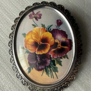 Pode incluir: Broche ovalado prateado com um design floral. O broche apresenta um buquê de amores-perfeitos em tons de amarelo, laranja e roxo, sobre um fundo claro. O broche tem uma borda ornamentada em estilo filigrana.