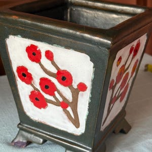 Può includere: Un vaso quadrato color bronzo scuro con quattro piedini. Ogni lato presenta un pannello bianco incassato decorato con un ramo marrone dipinto a mano e fiori rossi vivaci con centri neri. Il vaso ha un bordo superiore ampio e aperto.
