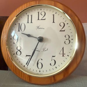 Puede incluir: Un reloj de pared redondo con un marco de madera de color marrón claro y un bisel dorado. La esfera es de color blanco roto con números arábigos negros, manecillas negras y el texto Hanson y Eden Forest en el centro. Made in Britain abajo.