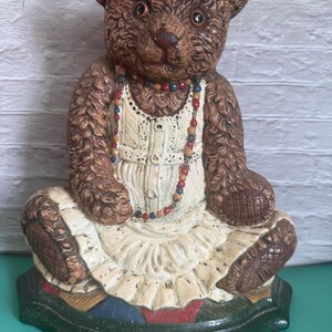Könnte beinhalten: Eine braune Bärenfigur mit weißem Kleid und bunter Perlenkette. Der Bär sitzt auf einem grünen Sockel mit Patchwork-Design. Die Figur hat ein Vintage- und rustikales Aussehen.