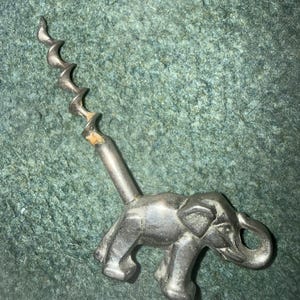 Könnte beinhalten: Ein silberfarbener Metallkorkenzieher mit einem Griff in Form einer skulpturalen Elefantenfigur. Der Korkenziehermechanismus erstreckt sich vom Rücken des Elefanten nach oben, wobei eine spiralförmige Metallspindel an einem zylindrischen Schaft befestigt ist.