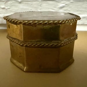 Pode incluir: Pequena caixa de latão antiga com formato octogonal. A caixa tem uma tampa articulada e é decorada com uma borda em forma de corda ao redor da parte superior e inferior. A caixa está sobre uma superfície marrom clara.