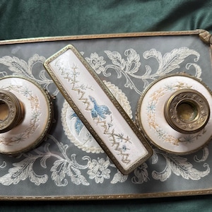 Op de afbeelding: Een antieke toilettafelset met een rechthoekige lade met bloemenborduurwerk, twee ronde containers en een rechthoekige borstel. De set heeft een goudkleurig metalen frame en accenten, met een lichtroze en blauw kleurenschema.