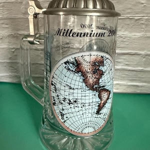 Könnte beinhalten: Klarer Bierkrug aus Glas mit einem Metallklappdeckel und Griff. Der Krug zeigt ein Weltkarten-Design und den Text "Millennium 2000". Das Glas hat einen dekorativen Boden.