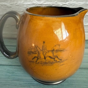 May include: A round ceramic jug with a glossy amber-coloured glaze and a fine crackle finish. The jug features a black line-art illustration of a person riding a horse across a field. It has a dark grey handle and a dark grey rim.