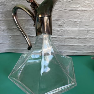 Può includere: Una caraffa di vetro trasparente con manico e beccuccio in metallo color argento. La caraffa ha un design geometrico sfaccettato e un coperchio incernierato. Il manico è curvo.