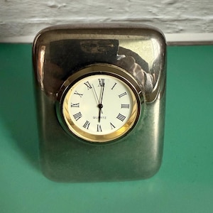 May include: A silver-toned quartz clock with a rectangular body and rounded edges. The clock face is white with black Roman numerals and gold-colored hands and a gold-colored bezel. The word "Quartz" is printed on the clock face.
