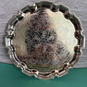 Può includere: Un vassoio da portata rotondo in metallo color argento con bordo smerlato e un intricato motivo floreale inciso e simmetrico al centro. La superficie riflettente presenta dettagliati motivi a volute e foglie che si irradiano da un punto centrale.