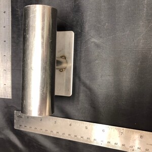 May include: A silver-colored metal rod holder with a rectangular mounting plate, positioned next to two metal rulers showing a length of approximately 6 inches for the tube and a width of approximately 3 inches for the mounting plate base.