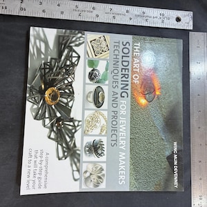 Puede incluir: Un libro titulado "The Art of Soldering for Jewelry Makers" con técnicas y proyectos. La portada presenta ejemplos de joyería y el nombre del autor, Wing Mun Devenney. Una regla muestra las medidas en centímetros.