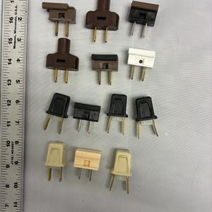 May include: A collection of vintage electrical plugs in various colors, including brown, black, and white. The plugs have two or three prongs and are arranged on a light gray surface. The image shows a variety of styles and designs.