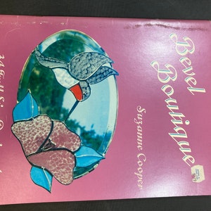 May include: A pink book titled "Bevel Boutique" by Suzanne Cooper. The cover features a stained glass design of a hummingbird and a flower.