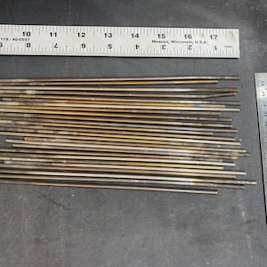 May include: A close-up of a pile of thin, gold-colored metal rods. The rods are arranged in a neat stack and are all approximately the same length. A ruler is visible in the background, indicating that the rods are about 16 inches long.
