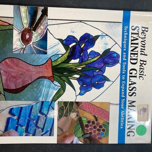 May include: A book titled "Beyond Basic Stained Glass Making" with a green sticker on the cover. The book cover features a stained glass design of a vase with purple irises and a close-up of a stained glass piece with a grape design.