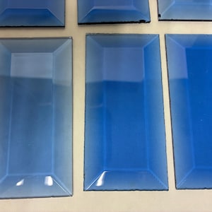 May include: A collection of rectangular, blue glass tiles. The tiles have a beveled edge and a gradient effect, transitioning from a lighter shade at the top to a deeper blue at the bottom. The tiles are arranged on a neutral surface.