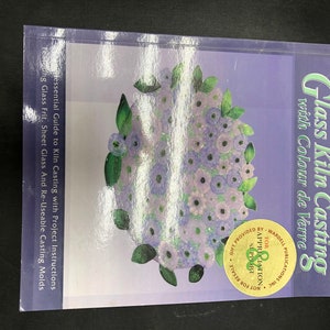 May include: A purple book titled "Glass Kiln Casting with Colour de Verre" by Jayne Persico. The cover features a floral design with a purple, white, and green color scheme. The book is an essential guide to kiln casting with project instructions, featuring glass frit, sheet glass, and reusable casting molds.