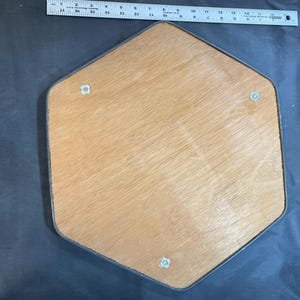 May include: A hexagonal wooden board with a dark brown border and three silver screws. The board is made of light wood with a natural grain pattern.