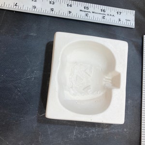 May include: White ceramic mold for making a pumpkin-shaped object. The mold has a raised design of a pumpkin face with a wide smile and two triangular eyes.