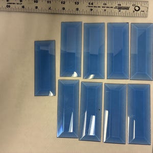 May include: A collection of eight rectangular, light blue glass tiles, with beveled edges, and a ruler for scale. The tiles vary in size, with the longest measuring approximately 5 inches. The tiles are arranged on a neutral surface.
